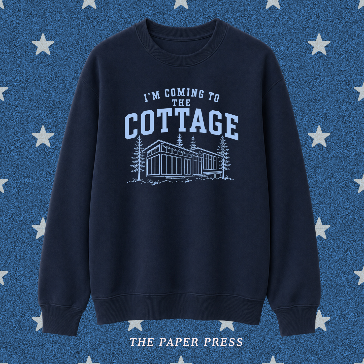 A navy blue crewneck sweatshirt with a light blue graphic print that reads 'I'M COMING TO THE COTTAGE' above an illustration of a cabin and trees. The sweatshirt is displayed against a textured blue background with white stars, and 'THE PAPER PRESS' is printed at the bottom.