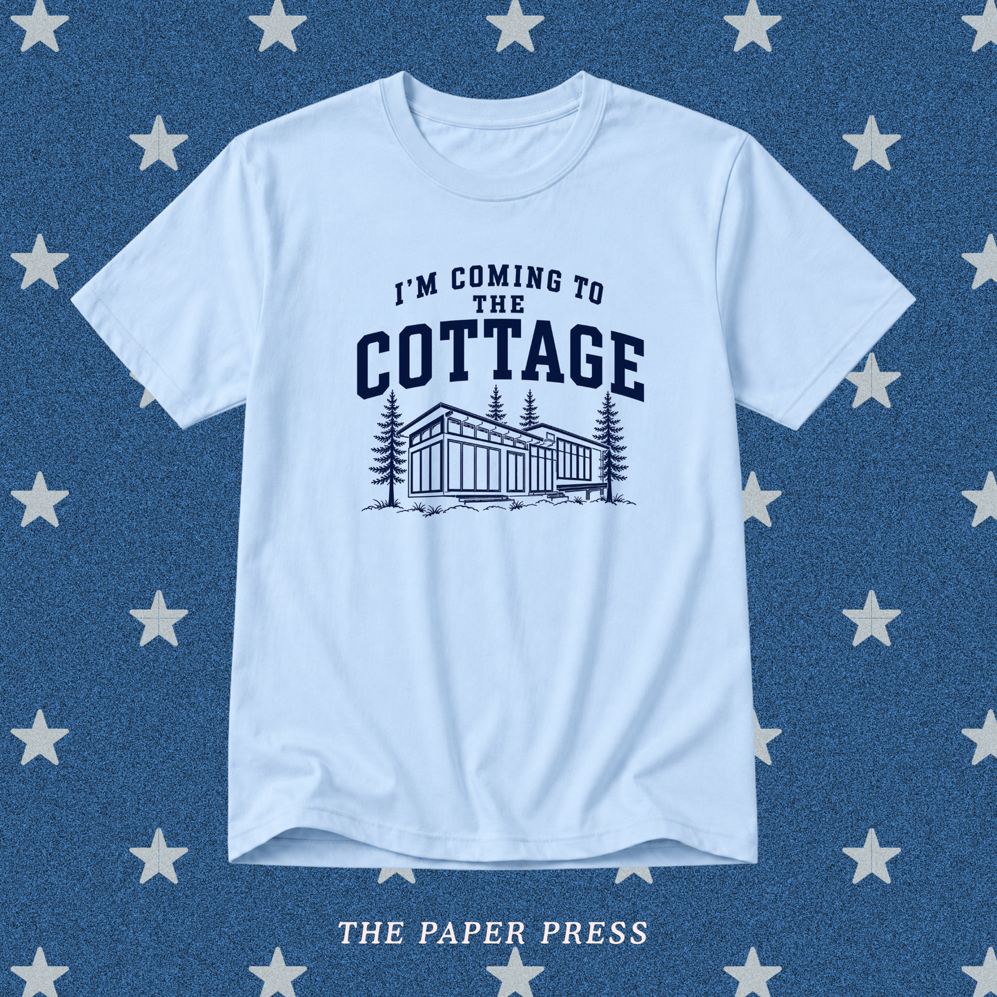 A light blue crewneck t-shirt with a dark navy graphic that reads 'I'M COMING TO THE COTTAGE' above an illustration of a cottage and pine trees. The shirt is displayed flat against a textured blue background patterned with white stars, with 'THE PAPER PRESS' printed at the bottom.