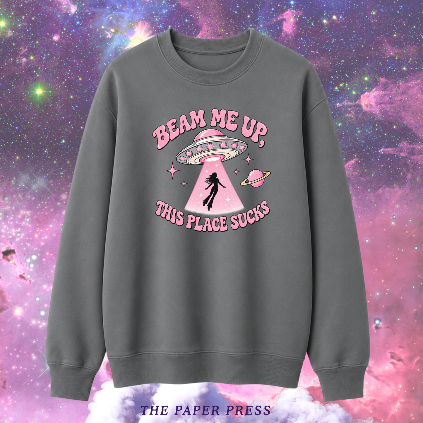 A dark gray crewneck sweatshirt featuring a pink graphic of a UFO, a person falling into a beam of light, and the text 'BEAM ME UP, THIS PLACE SUCKS'. The sweatshirt is displayed against a colorful pink and purple galaxy background, with 'THE PAPER PRESS' text at the bottom.