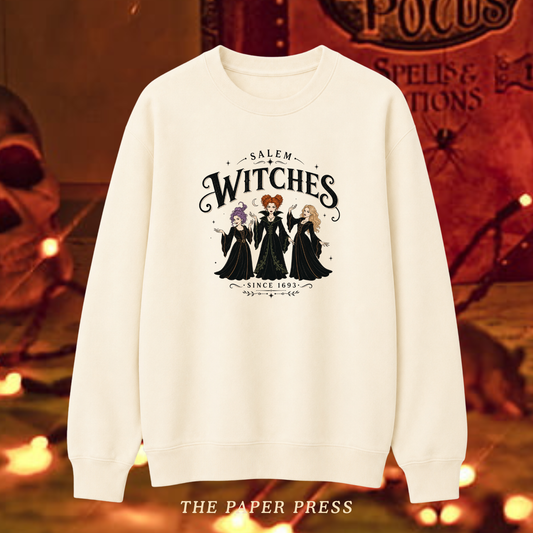 An off-white crewneck sweatshirt featuring a 'Salem Witches' graphic print with three witch characters and gothic text, displayed against a warm, spooky Halloween-themed background with the brand name 'THE PAPER PRESS' at the bottom.
