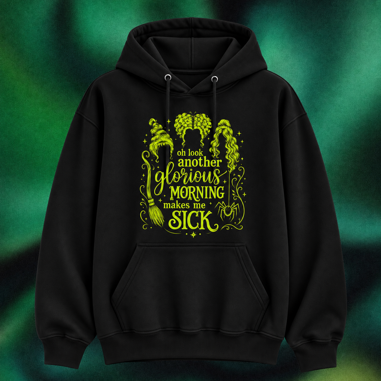 A black pullover hoodie with a vibrant lime green graphic on the front featuring the text 'oh look another glorious MORNING makes me SICK' surrounded by witchy illustrations like cauldrons, broomsticks, and a pentagram. The hoodie is displayed against a dark green, textured, smoky background.