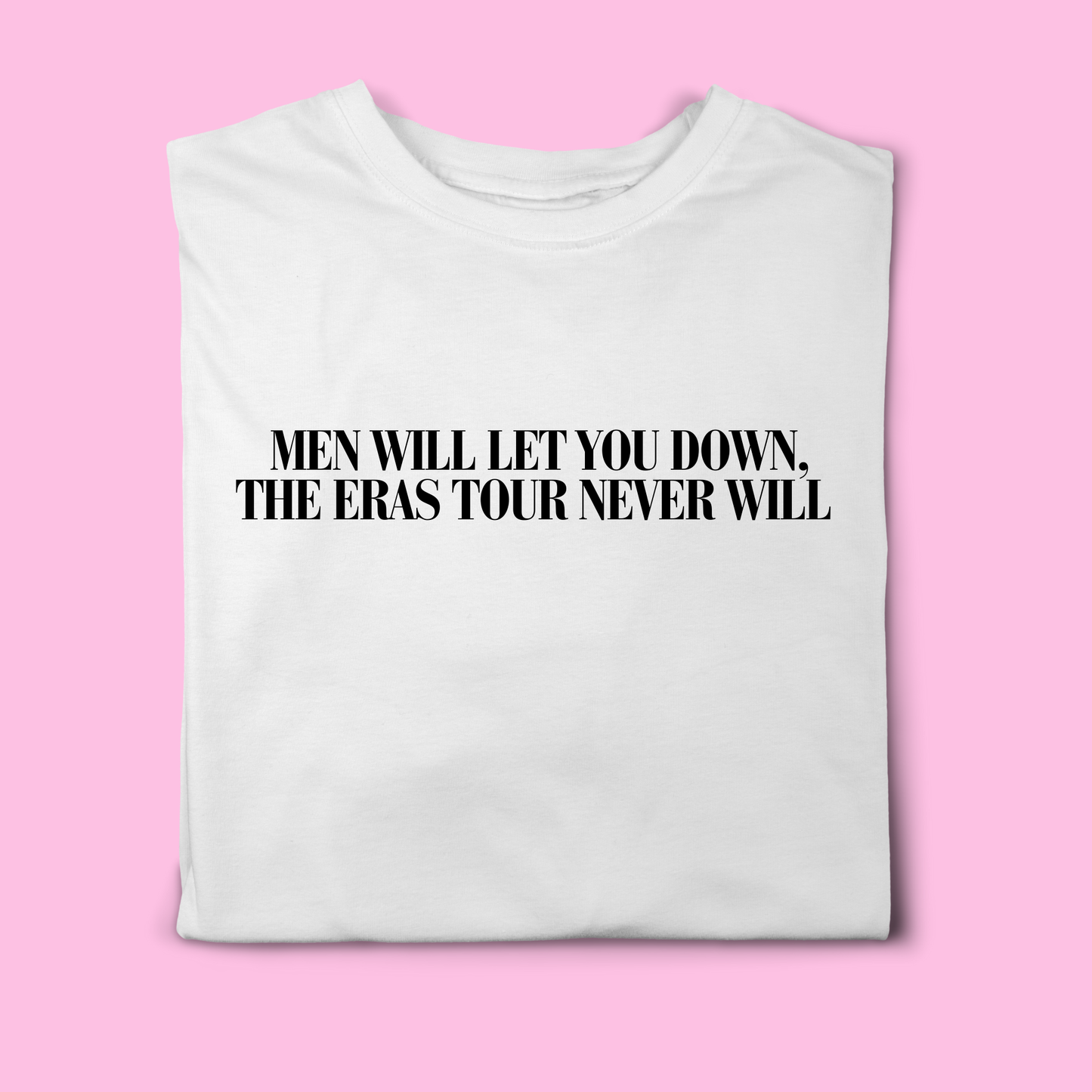 White t-shirt with black text reading “Men Will Let You Down, The Eras Tour Never Will”, folded flat on a pink background
