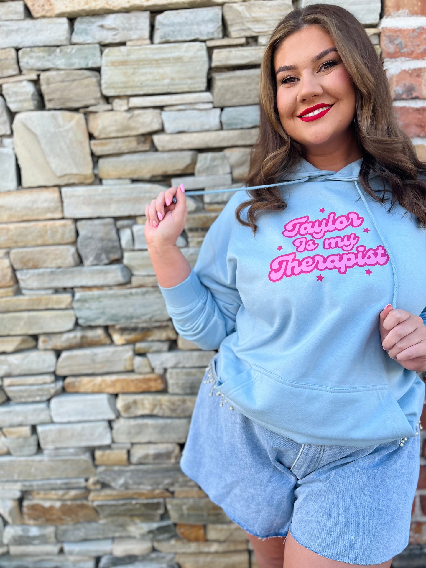A woman wearing a blue hoodie with the text 'TAYLOR IS MY THERAPIST' printed on the front, paired with denim shorts.