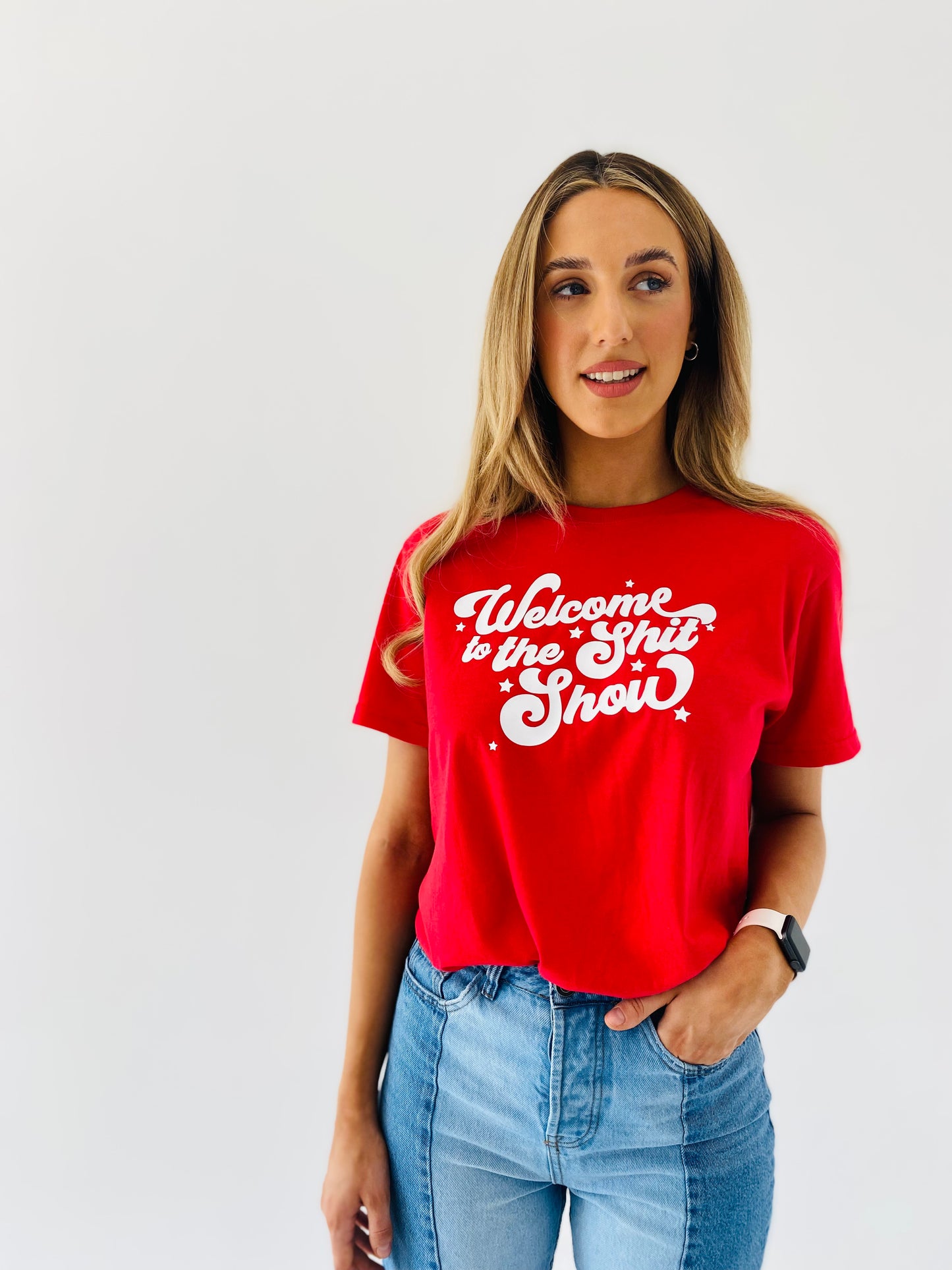 A woman wearing a red round neck t-shirt with the text 'WELCOME TO THE SHIT SHOW' in white capital letters.