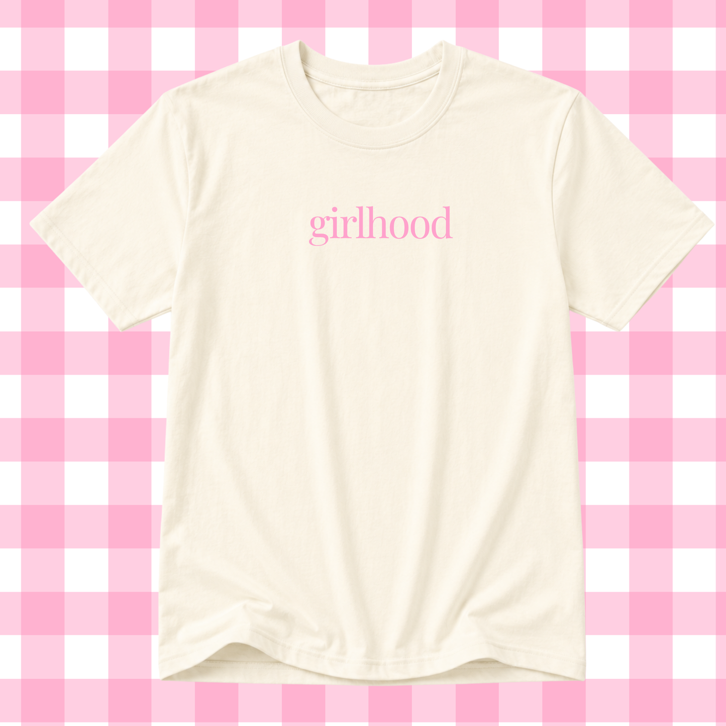 An off-white short-sleeve t-shirt with the word 'girlhood' printed in pink lowercase serif letters across the chest, displayed against a pink and white checkered background.