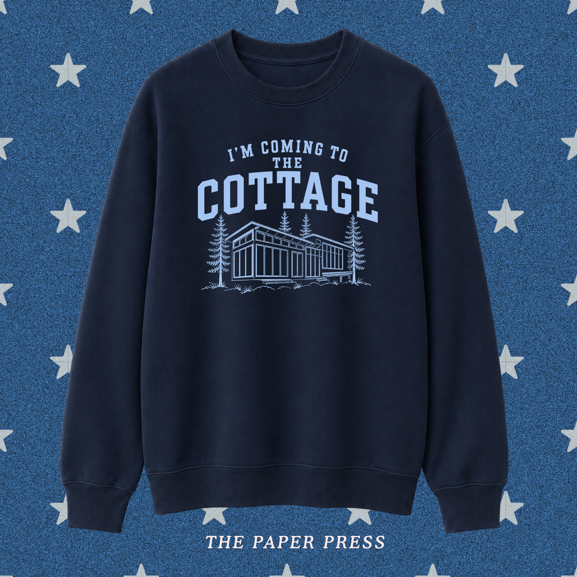 A navy blue crewneck sweatshirt with a light blue graphic print that reads 'I'M COMING TO THE COTTAGE' above an illustration of a cabin and trees. The sweatshirt is displayed against a textured blue background with white stars, and 'THE PAPER PRESS' is printed at the bottom.