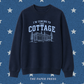 A navy blue crewneck sweatshirt with a light blue graphic print that reads 'I'M COMING TO THE COTTAGE' above an illustration of a cabin and trees. The sweatshirt is displayed against a textured blue background with white stars, and 'THE PAPER PRESS' is printed at the bottom.