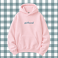 A light pink pullover hoodie with the word 'girlhood' printed in dark teal text on the chest, displayed against a blue and white gingham checkered background.