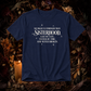 A navy blue short-sleeve graphic T-shirt laid flat on a dark autumn-themed background with dried leaves, pinecones, and a lit candle. The shirt has white text that reads, 'NO MAGIC IS STRONGER THAN SISTERHOOD.