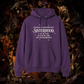 A purple pullover hoodie with a white printed quote on the chest, displayed against a dark background of dried flowers and foliage.