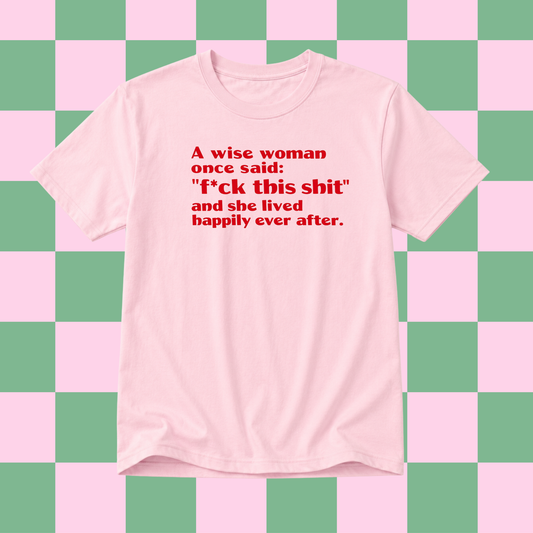 A light pink short-sleeve t-shirt with red printed text on the front, displayed against a pink and green checkered background.