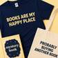 A navy blue graphic T-shirt with the phrase 'BOOKS ARE MY HAPPY PLACE' in yellow, shown alongside a cream canvas tote bag with the phrase 'PROBABLY BUYING ANOTHER BOOK' in navy, and a yellow book cover reading 'mystery book'.