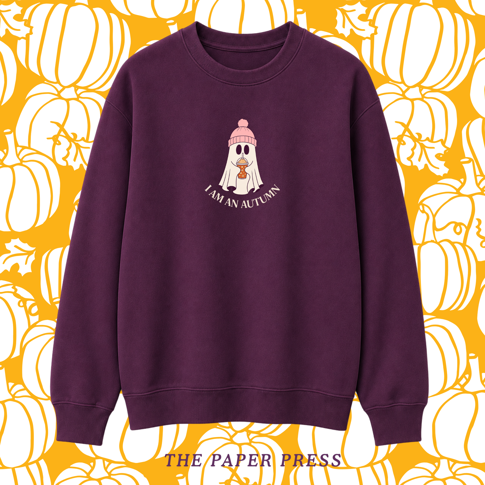 A dark purple crewneck sweatshirt featuring a cartoon ghost wearing a pink beanie with the text 'I AM AN AUTUMN' underneath. The sweatshirt is displayed against a yellow and white pumpkin patterned background with 'THE PAPER PRESS' text at the bottom.