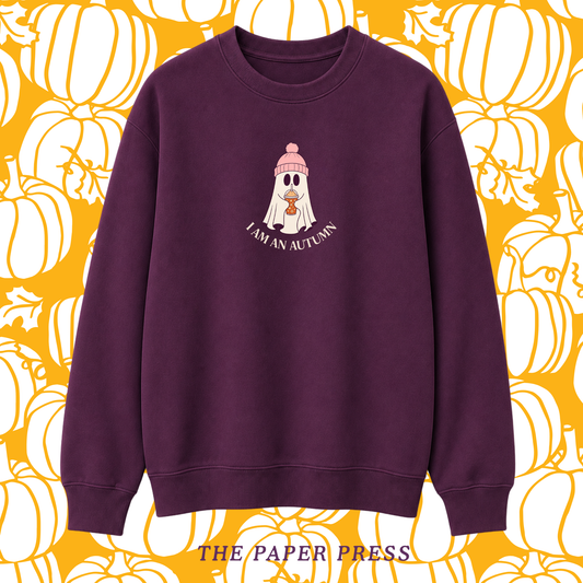 A dark purple crewneck sweatshirt featuring a cartoon ghost wearing a pink beanie with the text 'I AM AN AUTUMN' underneath. The sweatshirt is displayed against a yellow and white pumpkin patterned background with 'THE PAPER PRESS' text at the bottom.