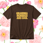 A dark brown short-sleeve t-shirt with the phrase 'GRANDMA HOBBIES ARE COOL' printed in bold yellow letters on the chest, presented against a white background decorated with pink and yellow watercolor flowers.