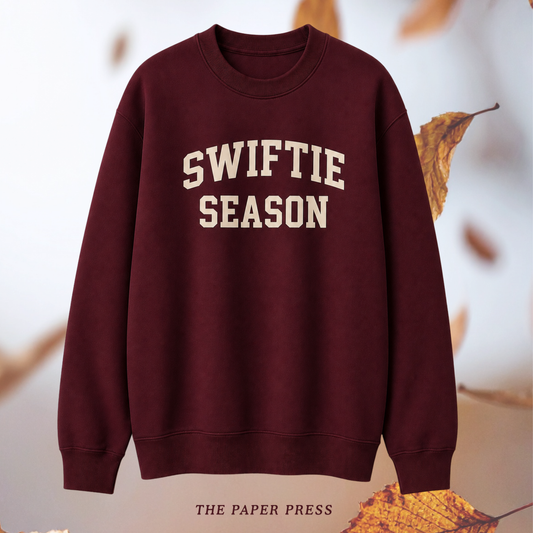 A crewneck sweatshirt in deep burgundy with 'SWIFTIE SEASON' printed in cream varsity-style lettering across the chest, displayed against a soft white background with scattered autumn leaves.