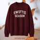 A crewneck sweatshirt in deep burgundy with 'SWIFTIE SEASON' printed in cream varsity-style lettering across the chest, displayed against a soft white background with scattered autumn leaves.