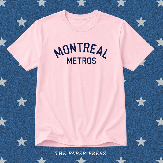 A light pink short-sleeve t-shirt with 'MONTREAL METROS' printed in navy blue arched text on the chest, displayed flat against a blue background patterned with white stars. 'THE PAPER PRESS' is printed at the bottom.