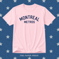 A light pink short-sleeve t-shirt with 'MONTREAL METROS' printed in navy blue arched text on the chest, displayed flat against a blue background patterned with white stars. 'THE PAPER PRESS' is printed at the bottom.