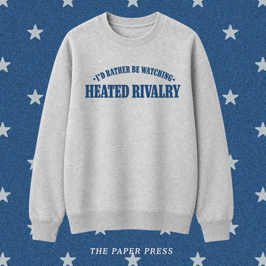 A light heather gray crewneck sweatshirt with blue text reading 'I'D RATHER BE WATCHING HEATED RIVALRY' on the chest, displayed against a blue background with white stars and the text 'THE PAPER PRESS' at the bottom.