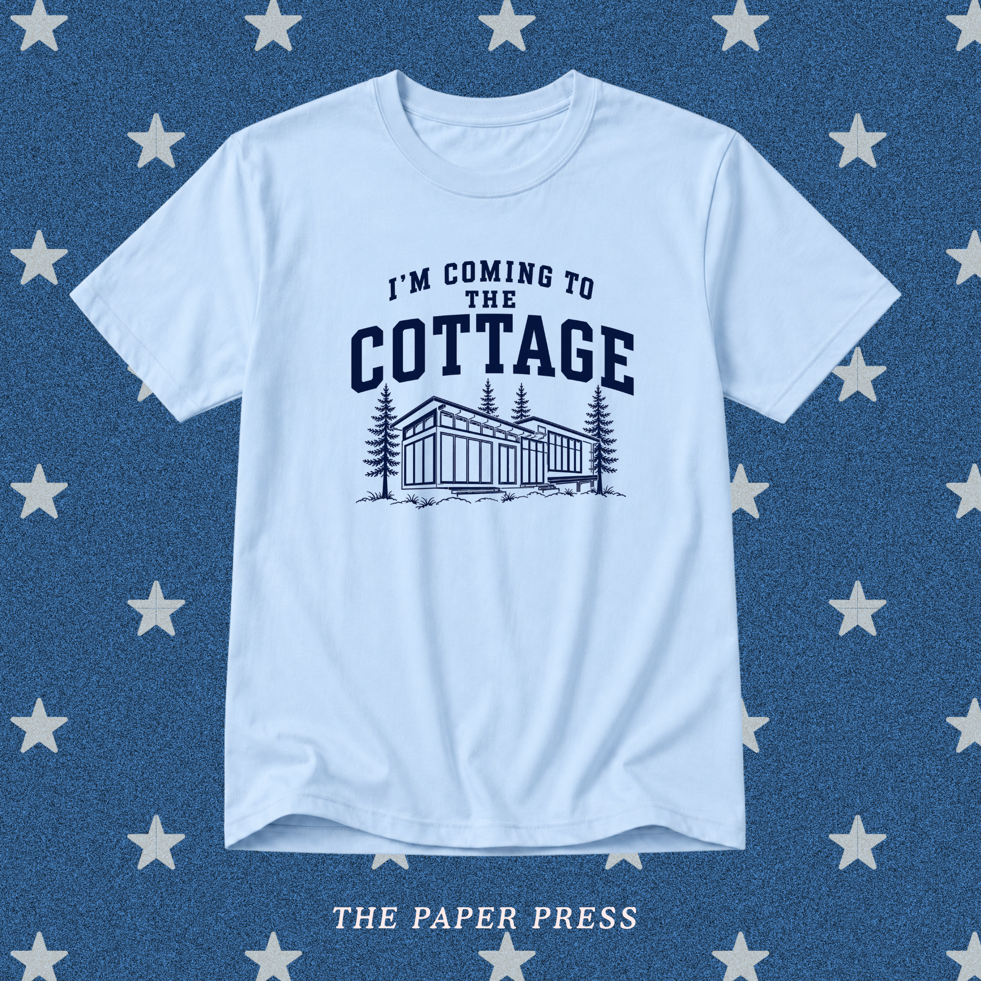A light blue crewneck t-shirt with a dark navy graphic that reads 'I'M COMING TO THE COTTAGE' above an illustration of a cottage and pine trees. The shirt is displayed flat against a textured blue background patterned with white stars, with 'THE PAPER PRESS' printed at the bottom.
