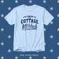 A light blue crewneck t-shirt with a dark navy graphic that reads 'I'M COMING TO THE COTTAGE' above an illustration of a cottage and pine trees. The shirt is displayed flat against a textured blue background patterned with white stars, with 'THE PAPER PRESS' printed at the bottom.