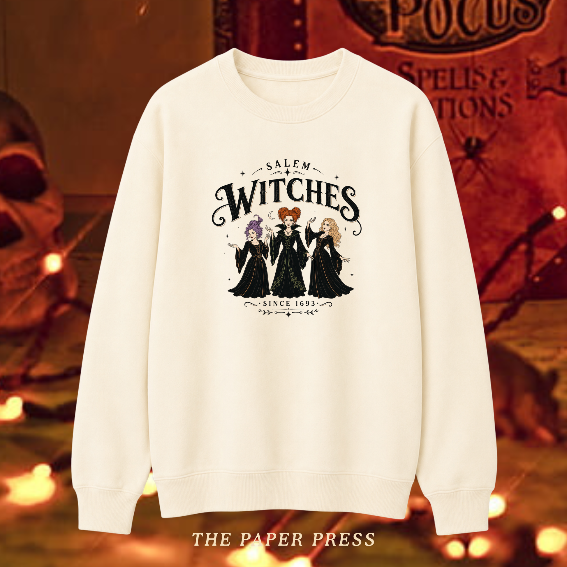 An off-white crewneck sweatshirt featuring a 'Salem Witches' graphic print with three witch characters and gothic text, displayed against a warm, spooky Halloween-themed background with the brand name 'THE PAPER PRESS' at the bottom.