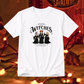 A white short-sleeve graphic t-shirt featuring the 'Salem Witches' illustration, displayed against a warm, blurred Halloween-themed background with orange lights and a cat.
