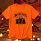 An orange short-sleeve graphic t-shirt featuring the characters 'Salem Witches' in black robes with colorful hair, displayed against a warm, blurred background with warm lights.