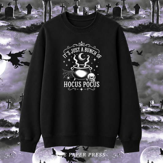 A black crewneck sweatshirt with a white graphic print reading 'IT'S JUST A BUNCH OF HOCUS POCUS' around an illustration of a cauldron, skull, and crescent moon. The sweatshirt is displayed against a spooky Halloween graveyard background with tombstones, crows, and full moons.