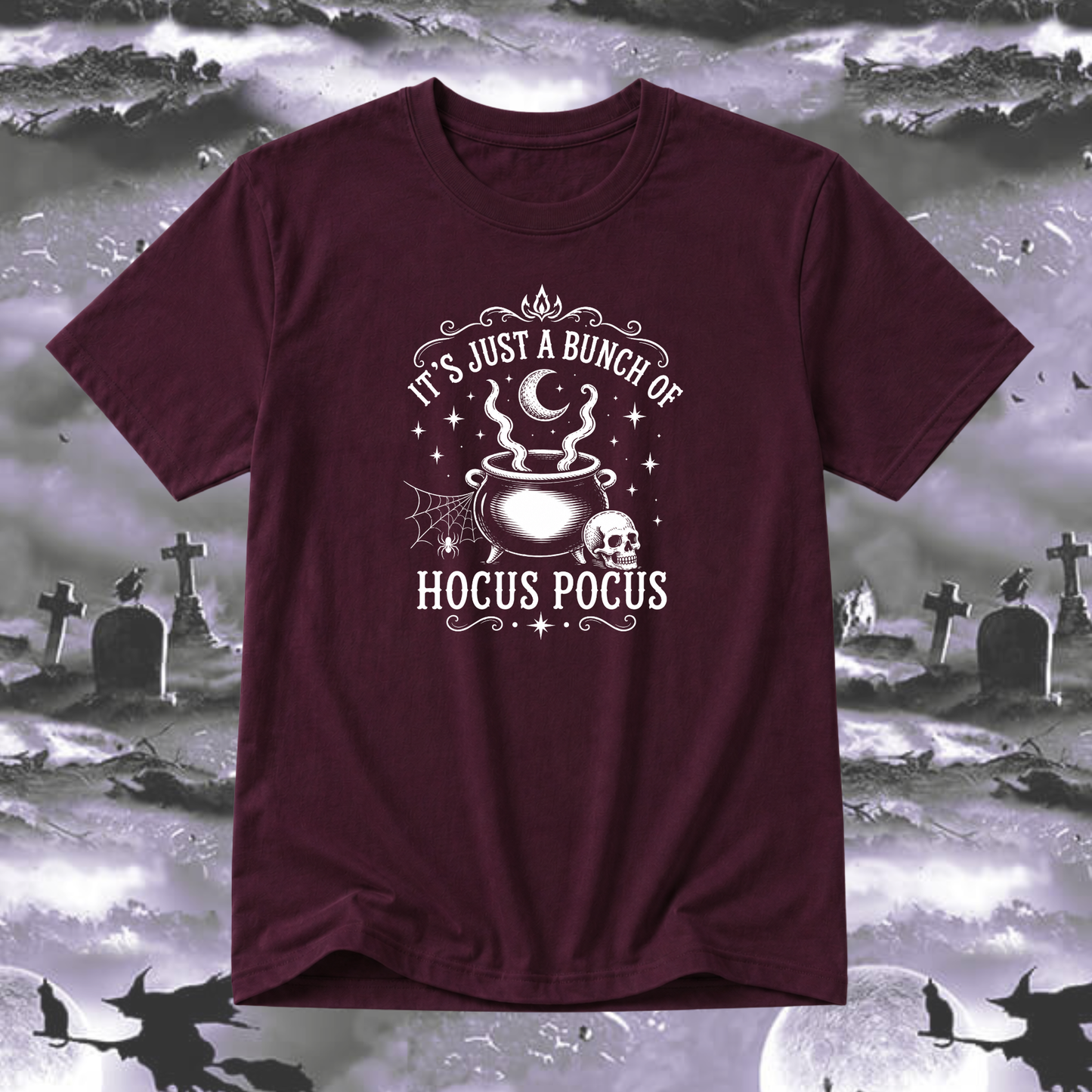 A maroon short-sleeve t-shirt with a white graphic print that reads 'IT'S JUST A BUNCH OF HOCUS POCUS' around an illustration of a cauldron, skull, spiderwebs, and stars. The shirt is displayed against a spooky graveyard background with silhouettes of bats and crosses.