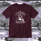A maroon short-sleeve t-shirt with a white graphic print that reads 'IT'S JUST A BUNCH OF HOCUS POCUS' around an illustration of a cauldron, skull, spiderwebs, and stars. The shirt is displayed against a spooky graveyard background with silhouettes of bats and crosses.