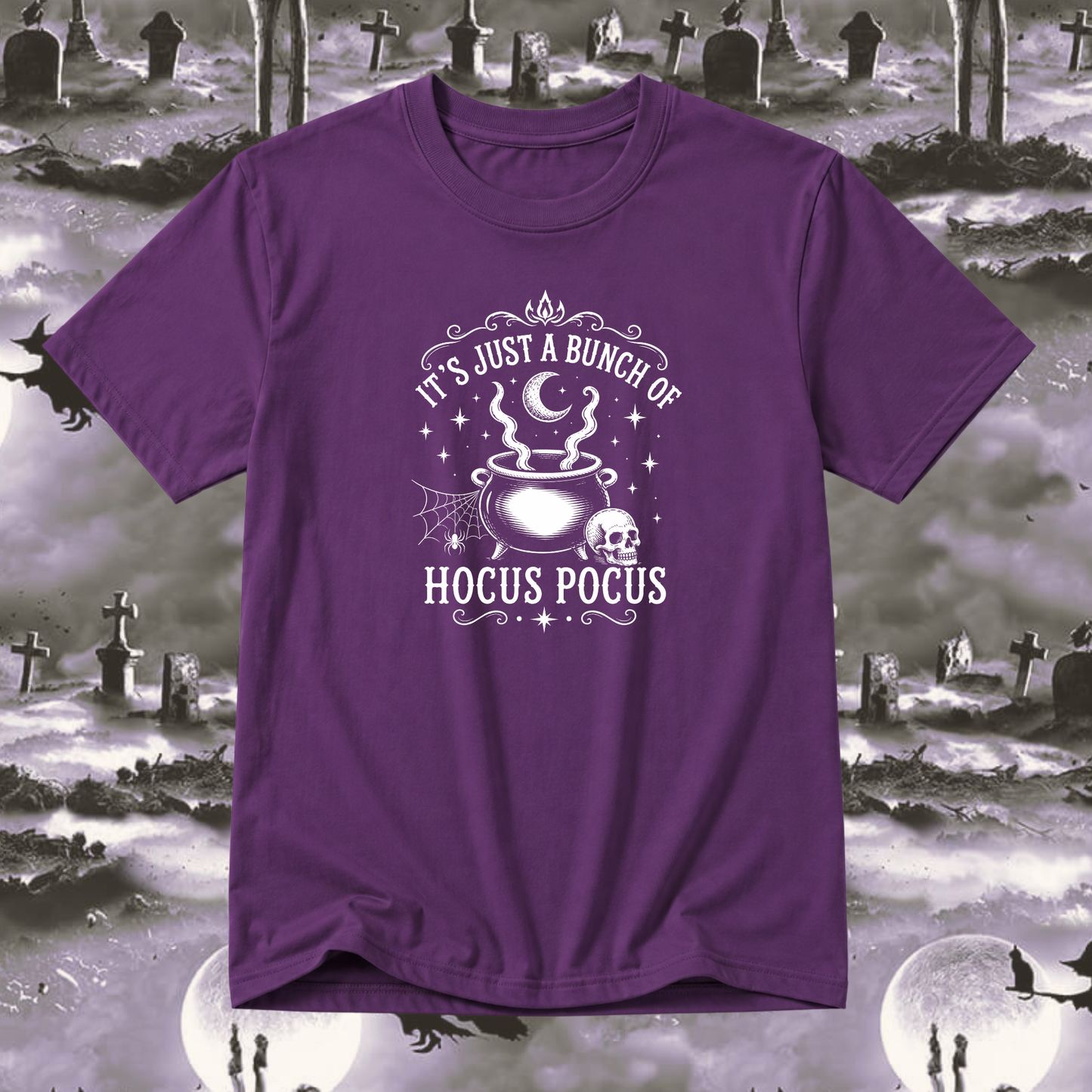 A purple short-sleeve t-shirt with a white graphic print that reads 'IT'S JUST A BUNCH OF HOCUS POCUS' around an illustration of a cauldron, skull, and stars. The shirt is displayed against a spooky, grayscale Halloween graveyard background with full moons and silhouettes.