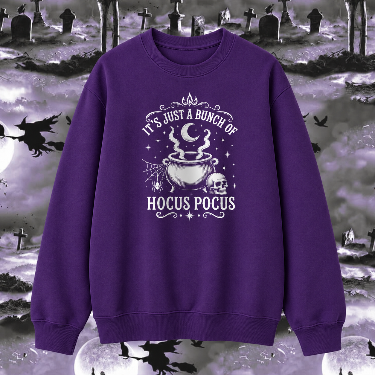A purple crewneck sweatshirt with a white graphic print reading 'IT'S JUST A BUNCH OF HOCUS POCUS' around an illustration of a cauldron, skull, and stars. The sweatshirt is displayed against a spooky Halloween-themed background of a graveyard with silhouettes of witches flying on broomsticks.