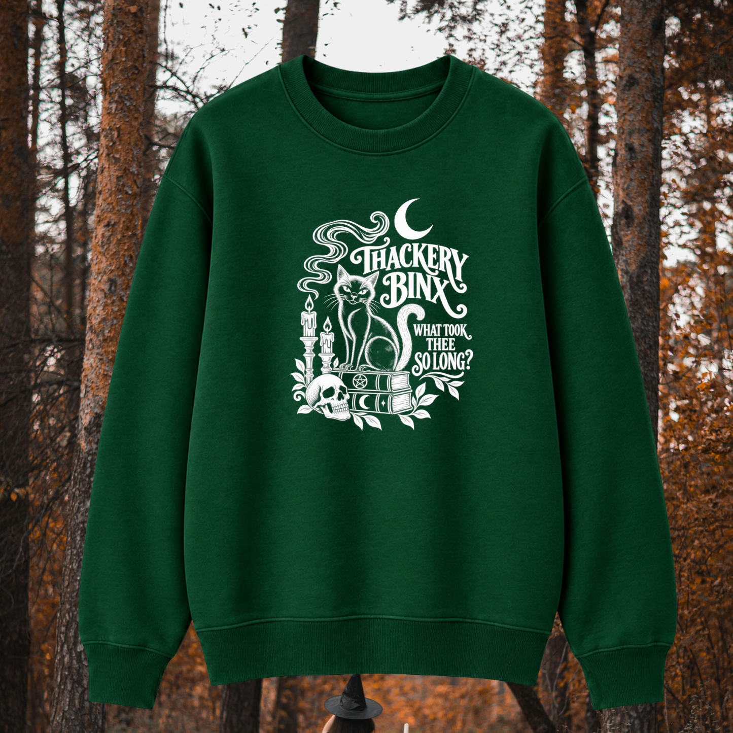 A dark green crewneck sweatshirt with a white graphic print featuring a cat, skull, and the text 'THACKERY BINX WHAT TOOL THEY SELLING?' displayed against a blurred autumn forest background.