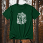 A dark green short-sleeve t-shirt with a white graphic print reading 'TRICKERY BINA' and 'WHAT TOOK, THEY SO LONG?' with a crescent moon, skull, candles, and books, displayed against a blurred autumn forest background.