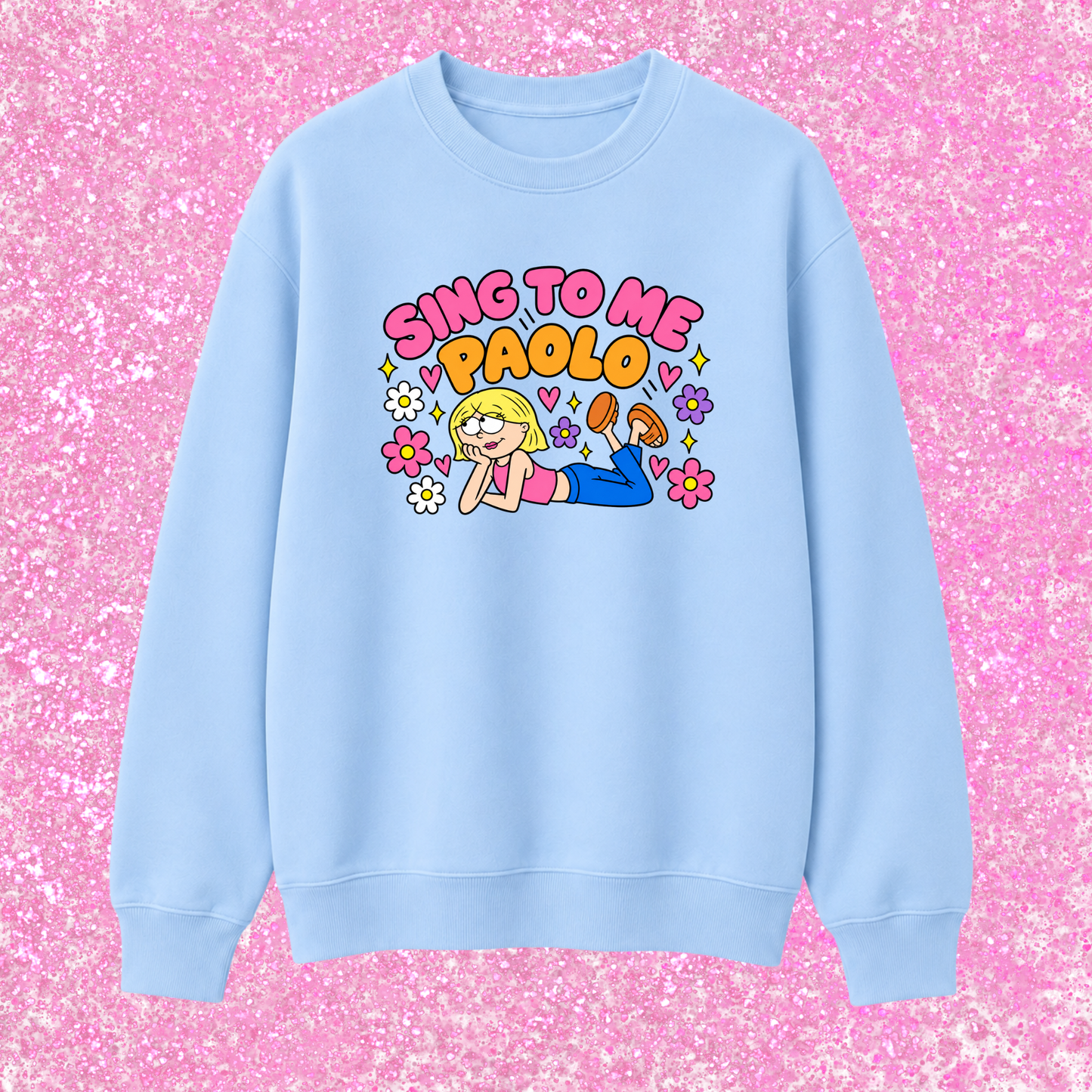 A light blue crewneck sweatshirt featuring a colorful cartoon graphic of a blonde girl lying down with the text 'SING TO ME PAOLO' in pink and orange bubble letters, surrounded by small flowers. The sweatshirt is displayed flat against a bright pink glittery background.