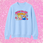 A light blue crewneck sweatshirt featuring a colorful cartoon graphic of a blonde girl lying down with the text 'SING TO ME PAOLO' in pink and orange bubble letters, surrounded by small flowers. The sweatshirt is displayed flat against a bright pink glittery background.
